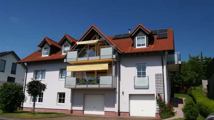 Helles Mehrfamilienhaus mit roten Dächern und Balkonen. Grüner Garten und blauer Himmel.