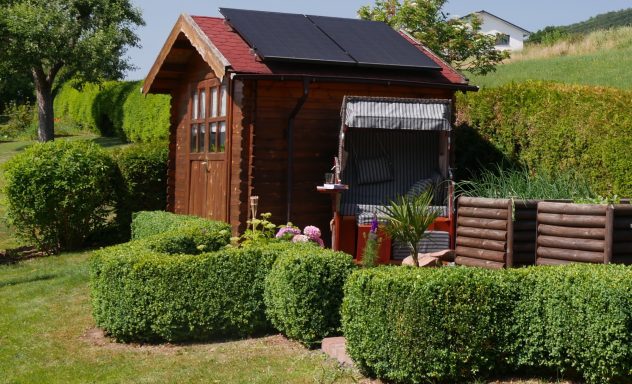Hölzernes Gartenhaus mit Solarpanel, umgeben von gepflegten Sträuchern und Pflanzen.