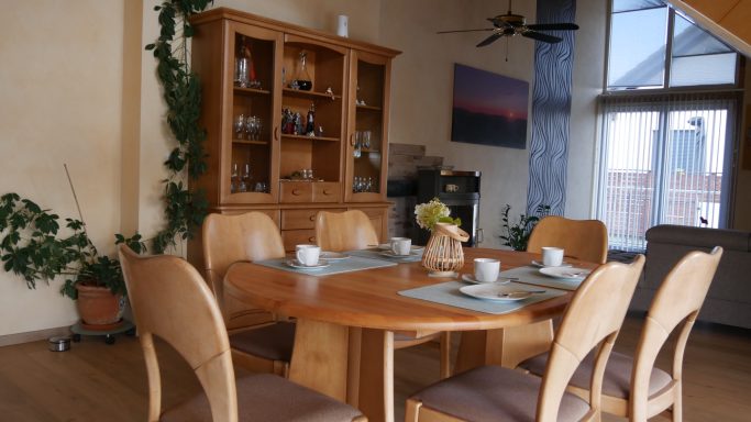 Holztisch mit sechs Stühlen in einem hellen Esszimmer, dekoriert mit Pflanzen und einem Schrank.