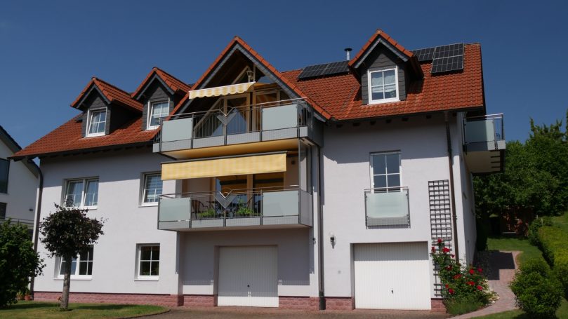 Helles, modernes Wohnhaus mit Balkon und Garagentor, umgeben von Grünflächen.