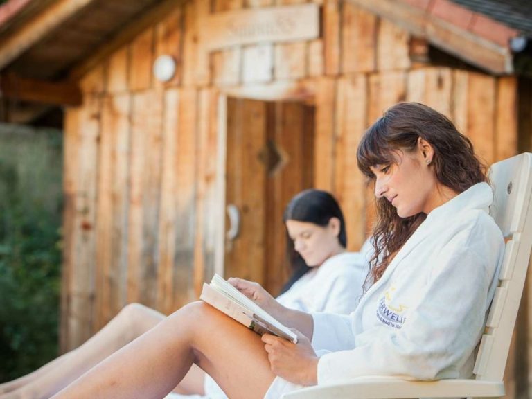 Zwei Frauen in Bademänteln entspannen und lesen vor einer Holzhausfassade.