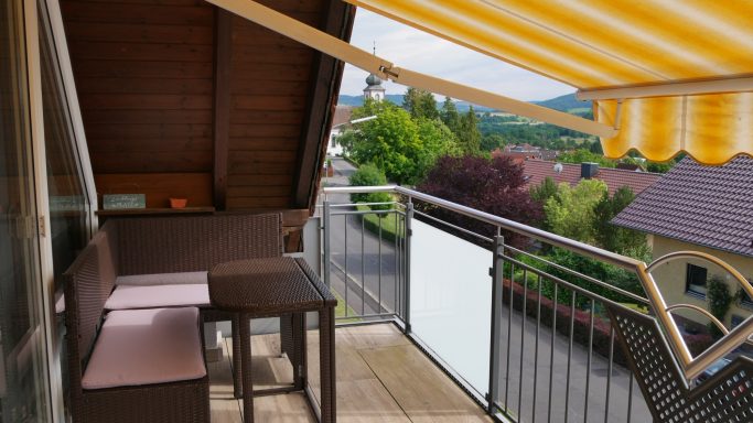 Balkon mit Holzgeplank, Tisch und Stühlen, gelb-weißem Sonnenschirm und grüner Umgebung.